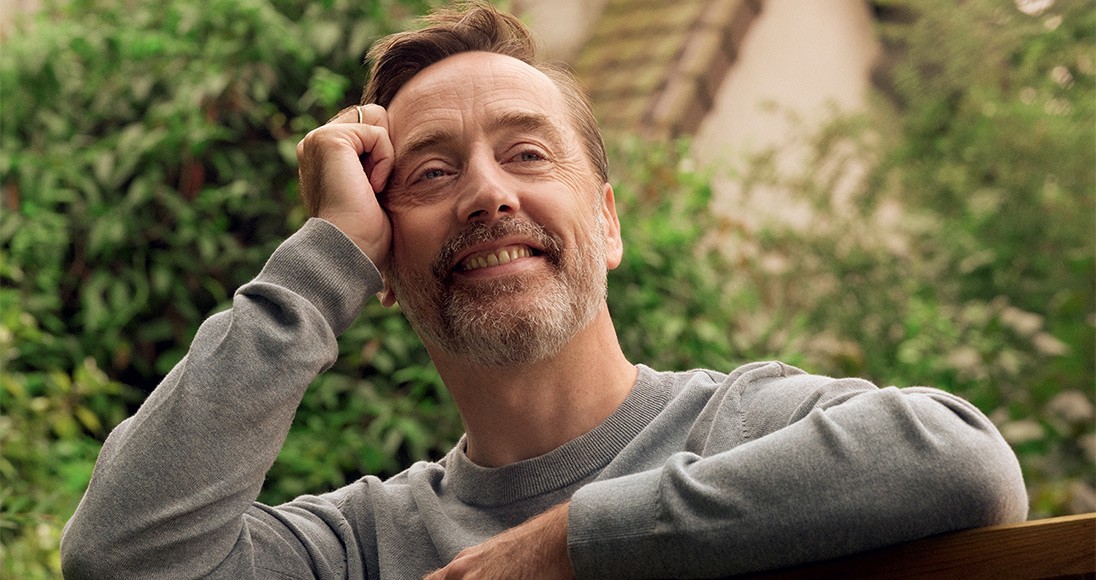 Un homme s'appuie à la barrière d'un jardin. Il a le sourire.